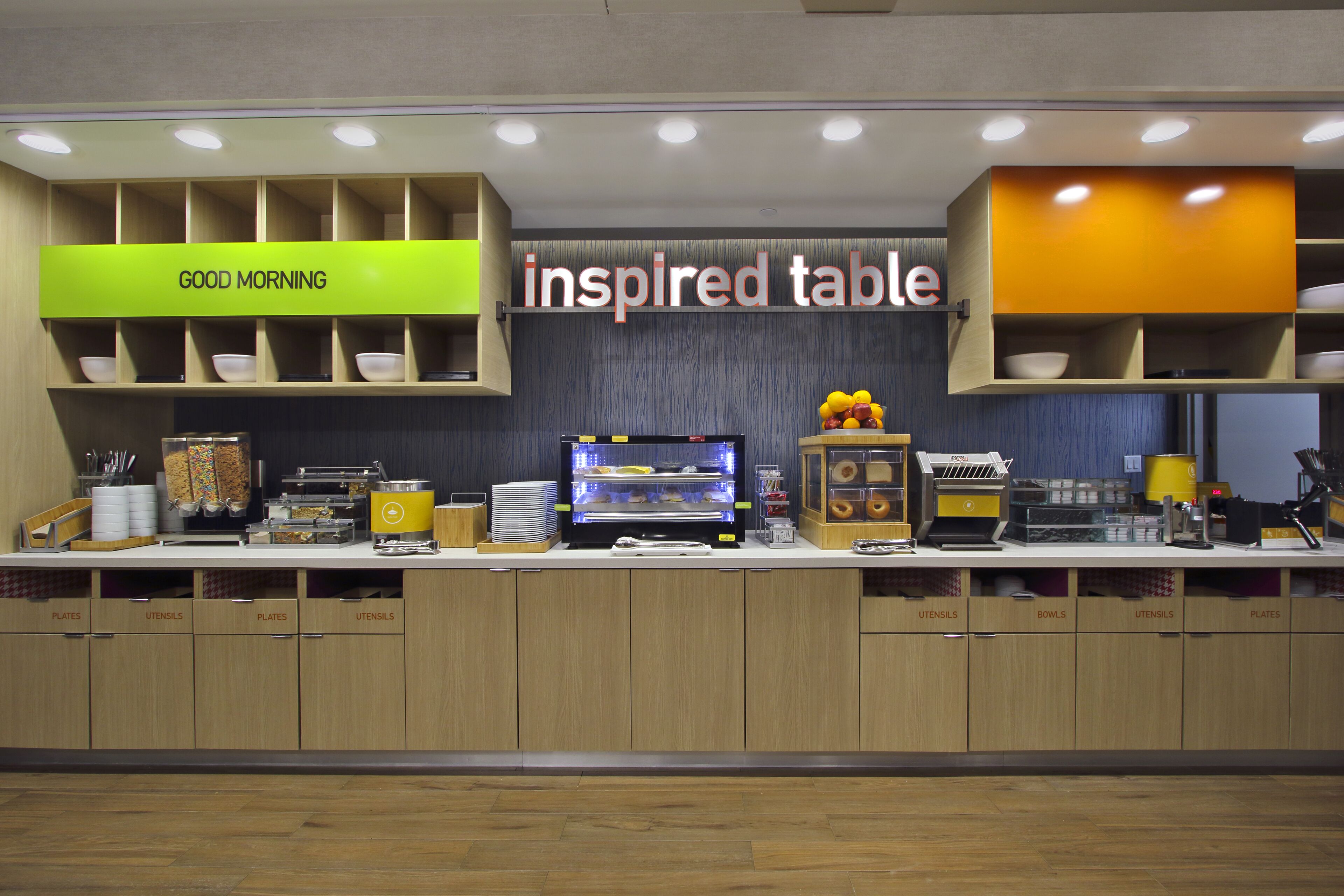 A breakfast bar with labeled storage for plates and utensils, a coffee machine, and a sign reading "inspired table".