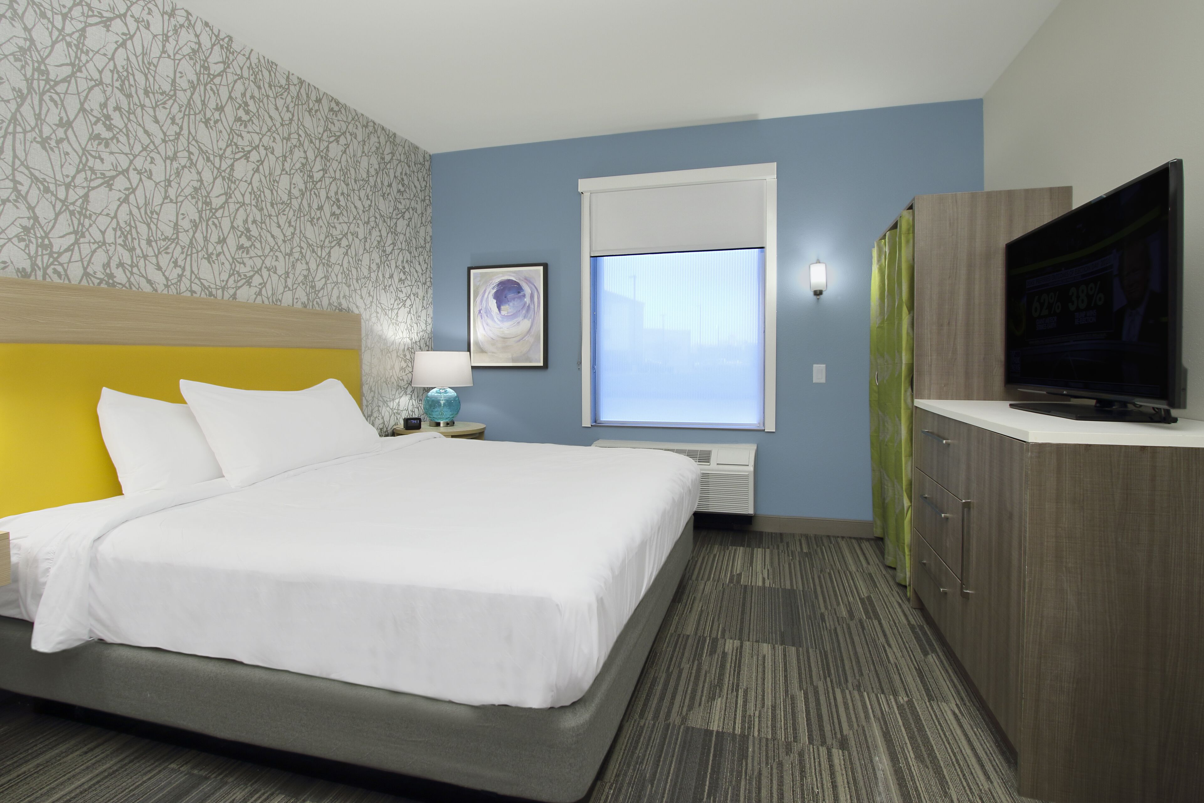 A hotel room with a bed, a television, a window with blinds, a nightstand with a lamp, and a wall with a patterned wallpaper.