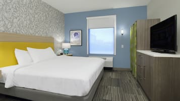 Habitación de hotel con cama, televisor, ventana con persianas, mesita de noche con lámpara y pared con papel tapiz estampado.