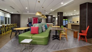 A modern lounge area with a green sofa, yellow chairs, a wooden table, and a reception desk. - Home2 Suites by Hilton Beaumont (Beaumont)
