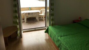 A room with a green bed, a small table with a chair outside, and a view of a body of water. - Apartments Tarcizio (Cres)