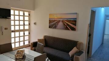 Un salon avec un canapé, une table sur laquelle se trouve un plateau, et un tableau représentant un coucher de soleil sur une plage.