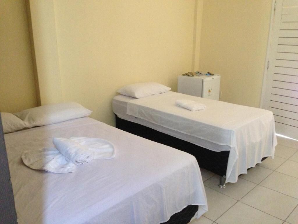 Dos camas individuales con sábanas blancas, un mueble pequeño y piso de baldosas en una habitación de hotel.
