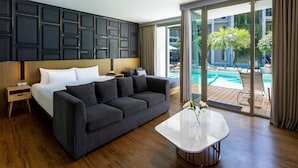 Habitación de hotel moderna con cama, sofá gris oscuro, mesa de centro blanca y vista a una piscina al aire libre.