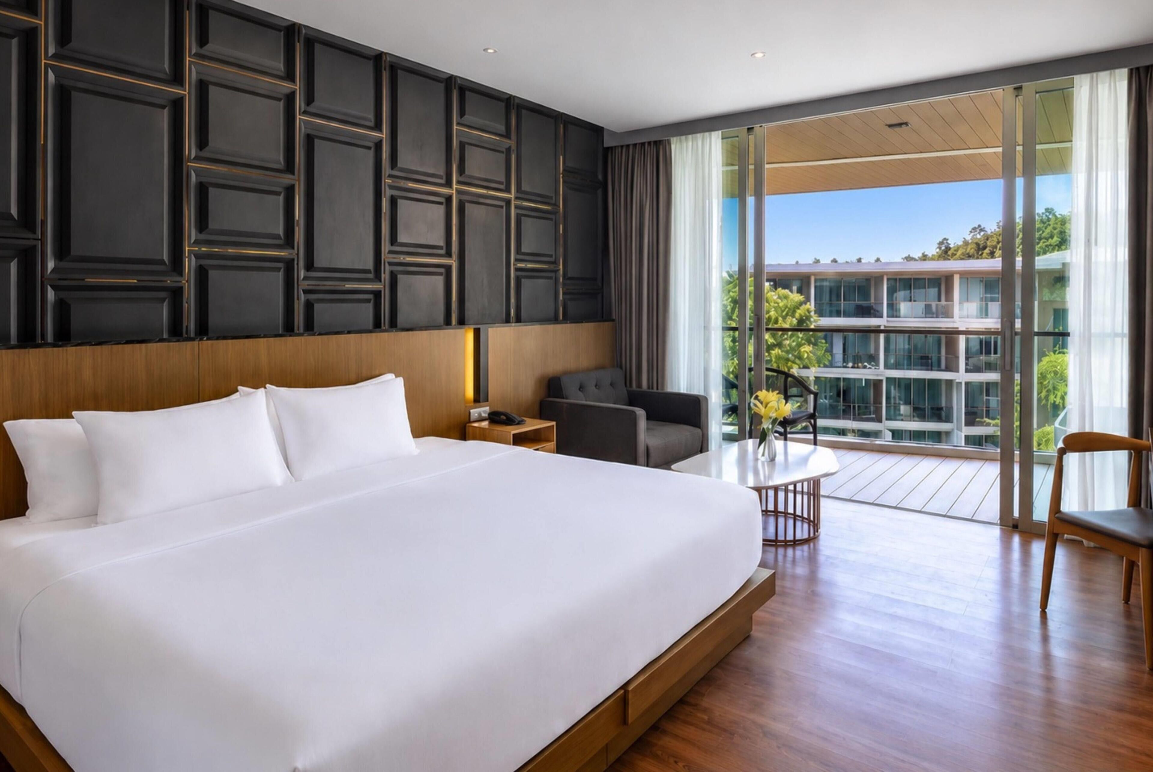 A large bed with white linens, a wooden headboard, a nightstand, a desk with a chair, and a view of a building through large windows.