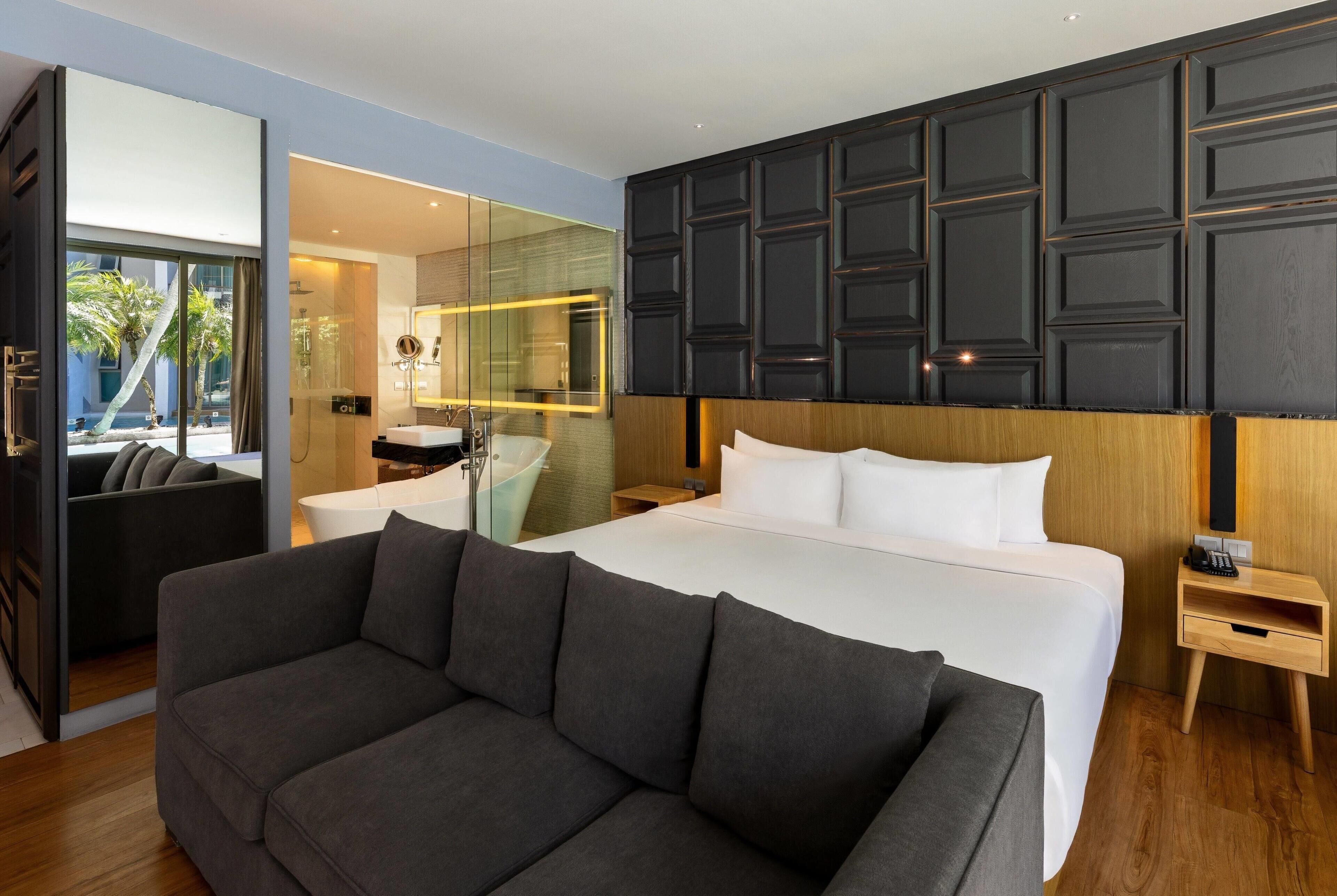 A modern hotel room with a large bed, a sofa, a glass shower enclosure, and a wooden headboard.