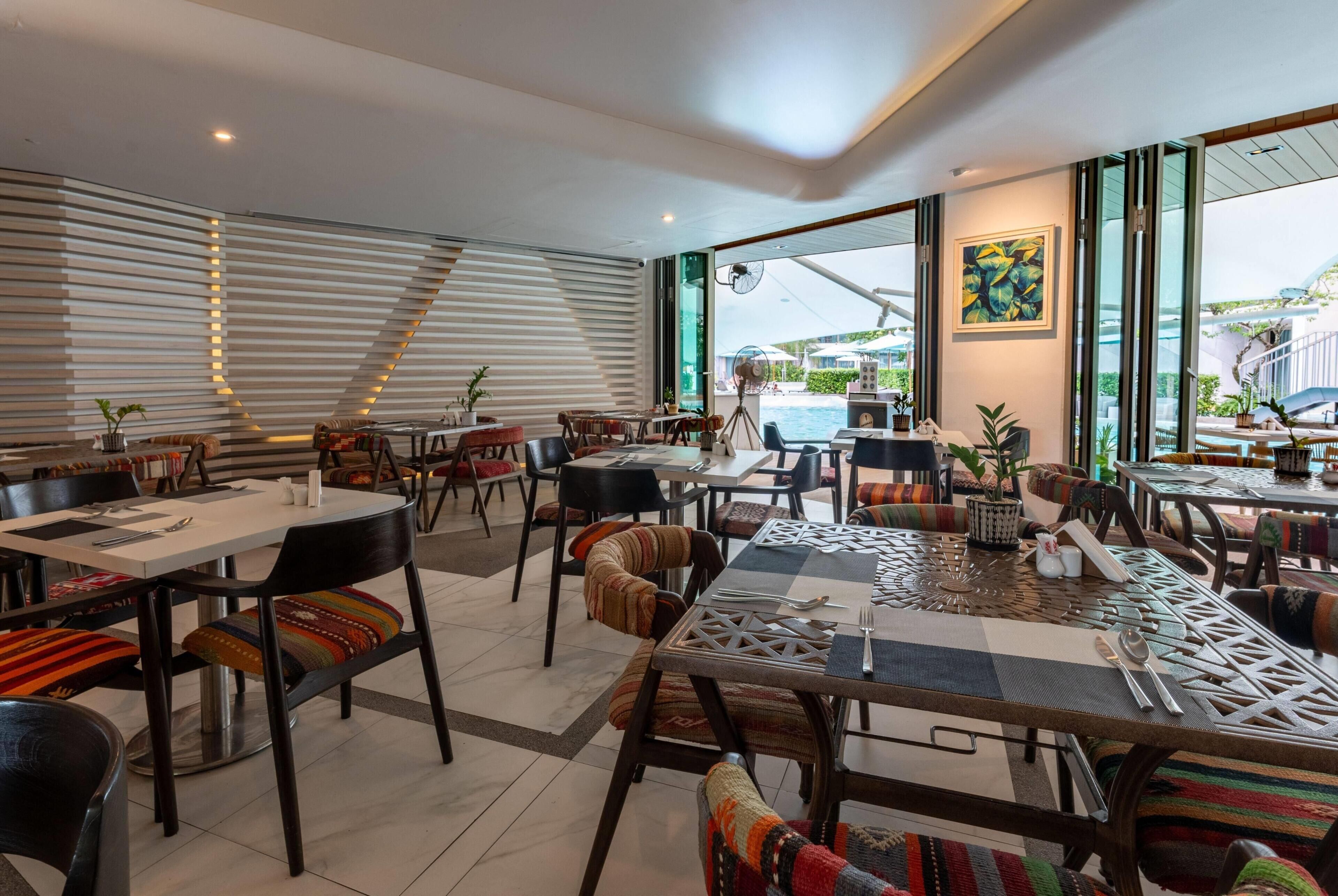 A modern dining area with multiple tables and chairs, large windows with a view of the outdoors, and a staircase leading to an upper level.