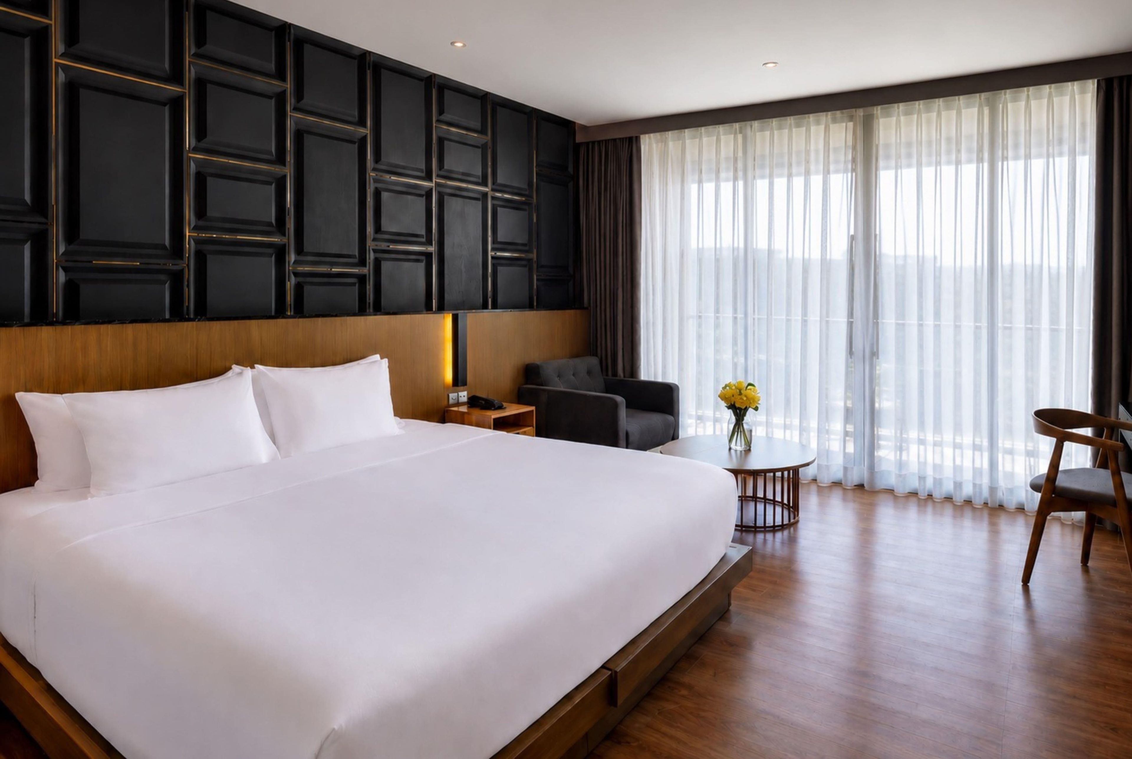 Habitació d'hotel amb un llit gran, una taula de treball, una cadira i vistes a l'exterior a través de cortines translúcides.