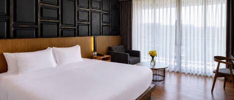 A hotel room with a large bed, a desk, a chair, and a view of the outdoors through sheer curtains.