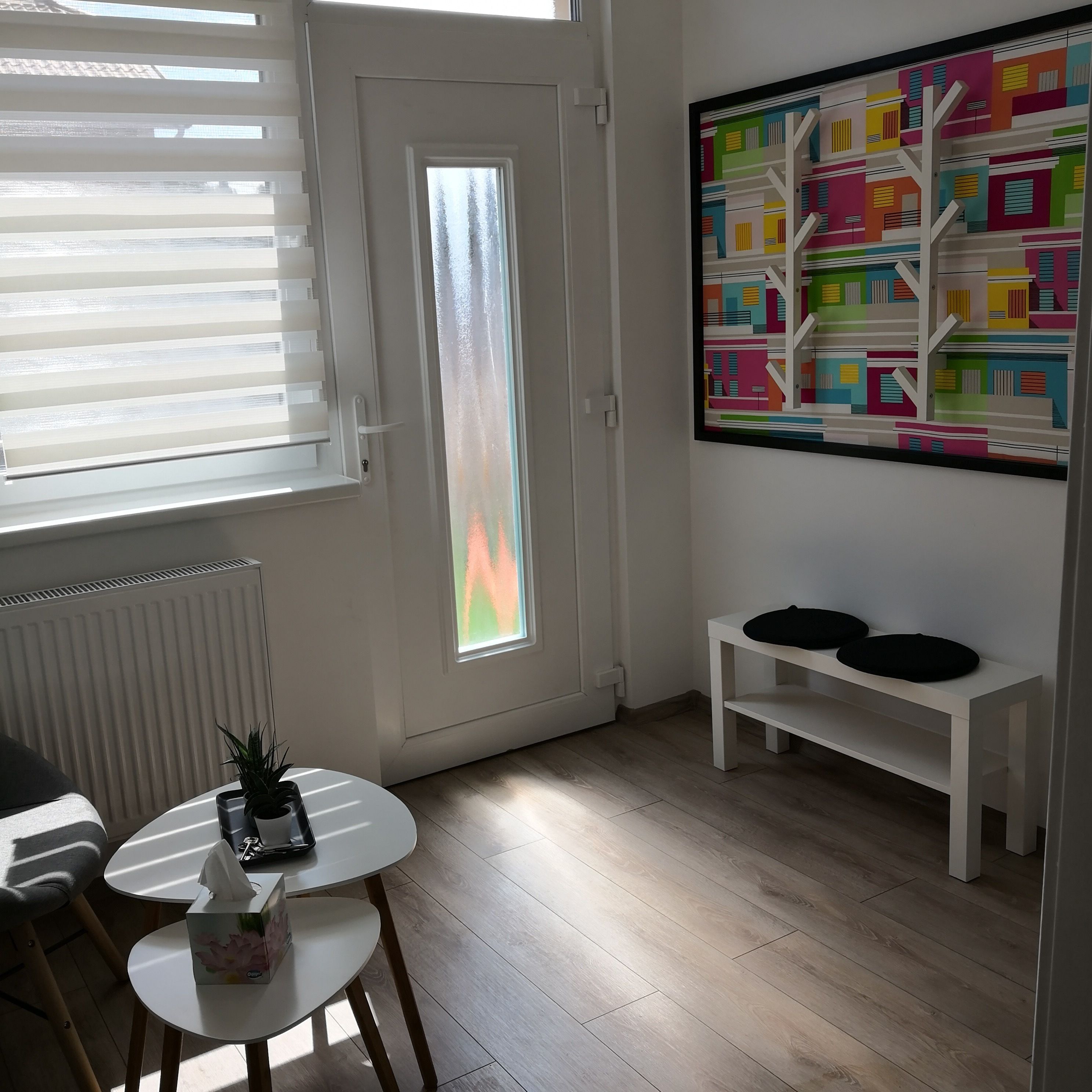 A room with a white door, a window with blinds, a radiator, a round table with a tissue box, a small potted plant, and a colorful abstract painting on the wall.