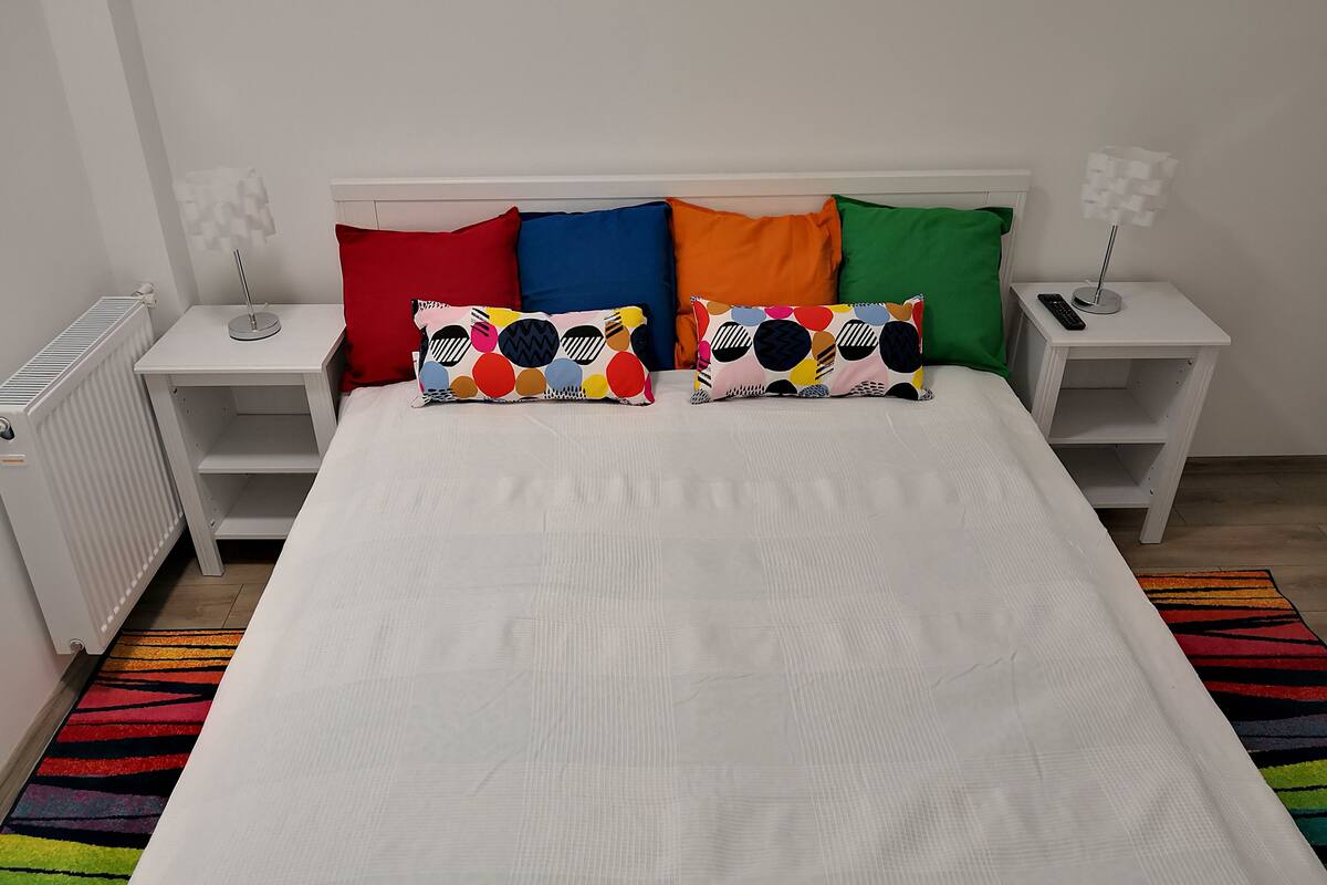 A bed with a white sheet, two pillows with colorful patterns, and a striped rug. There are two bedside tables with lamps and a radiator.