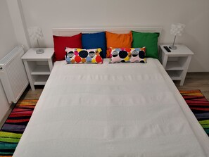 A bed with a white sheet, two pillows with colorful patterns, and a striped rug. There are two bedside tables with lamps and a radiator. - Green Garden Plus Apartman (Budapest)