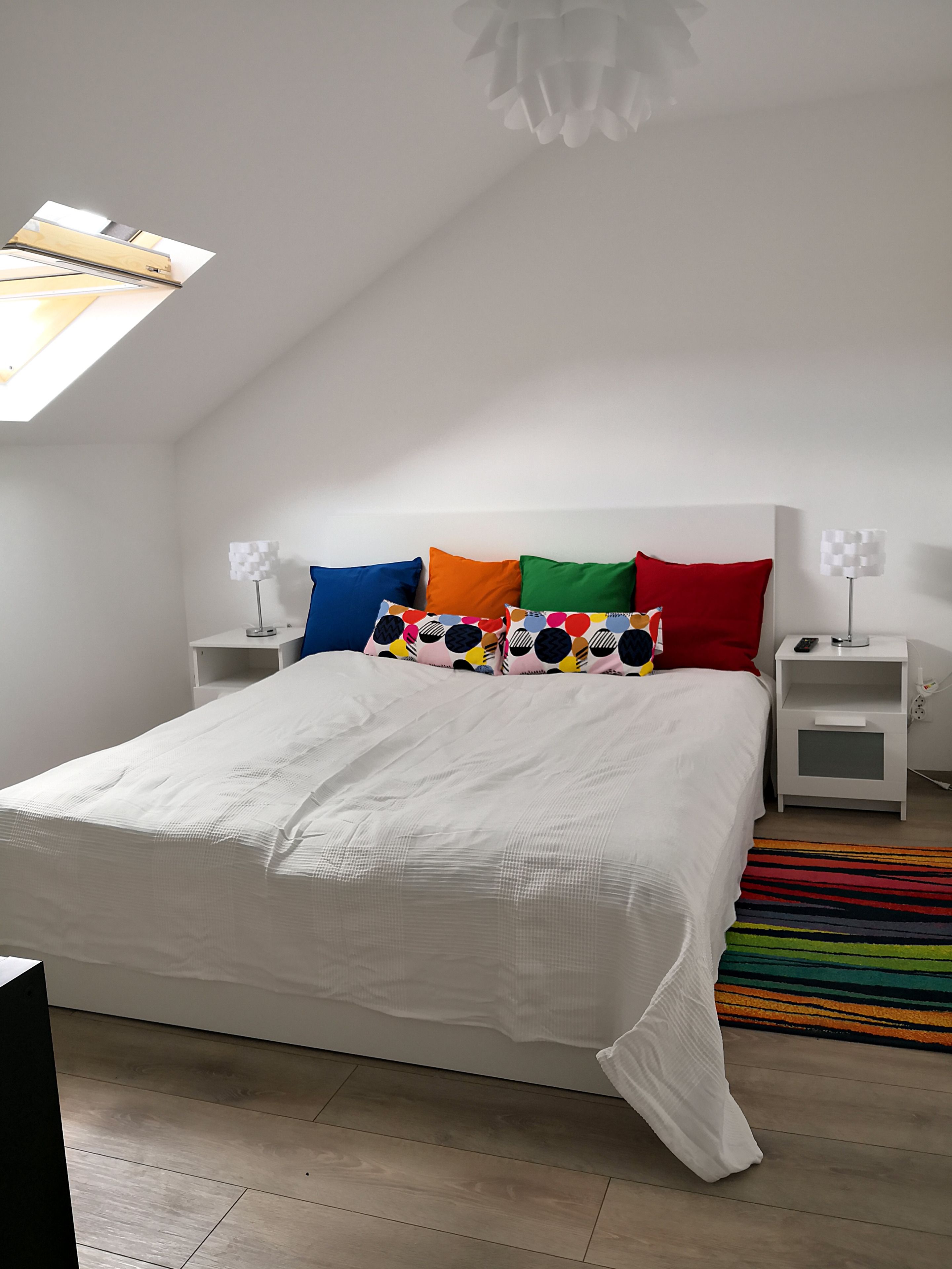 A modern bedroom with a sloped ceiling, a large bed with white bedding, colorful pillows, two bedside tables with lamps, and a striped rug.