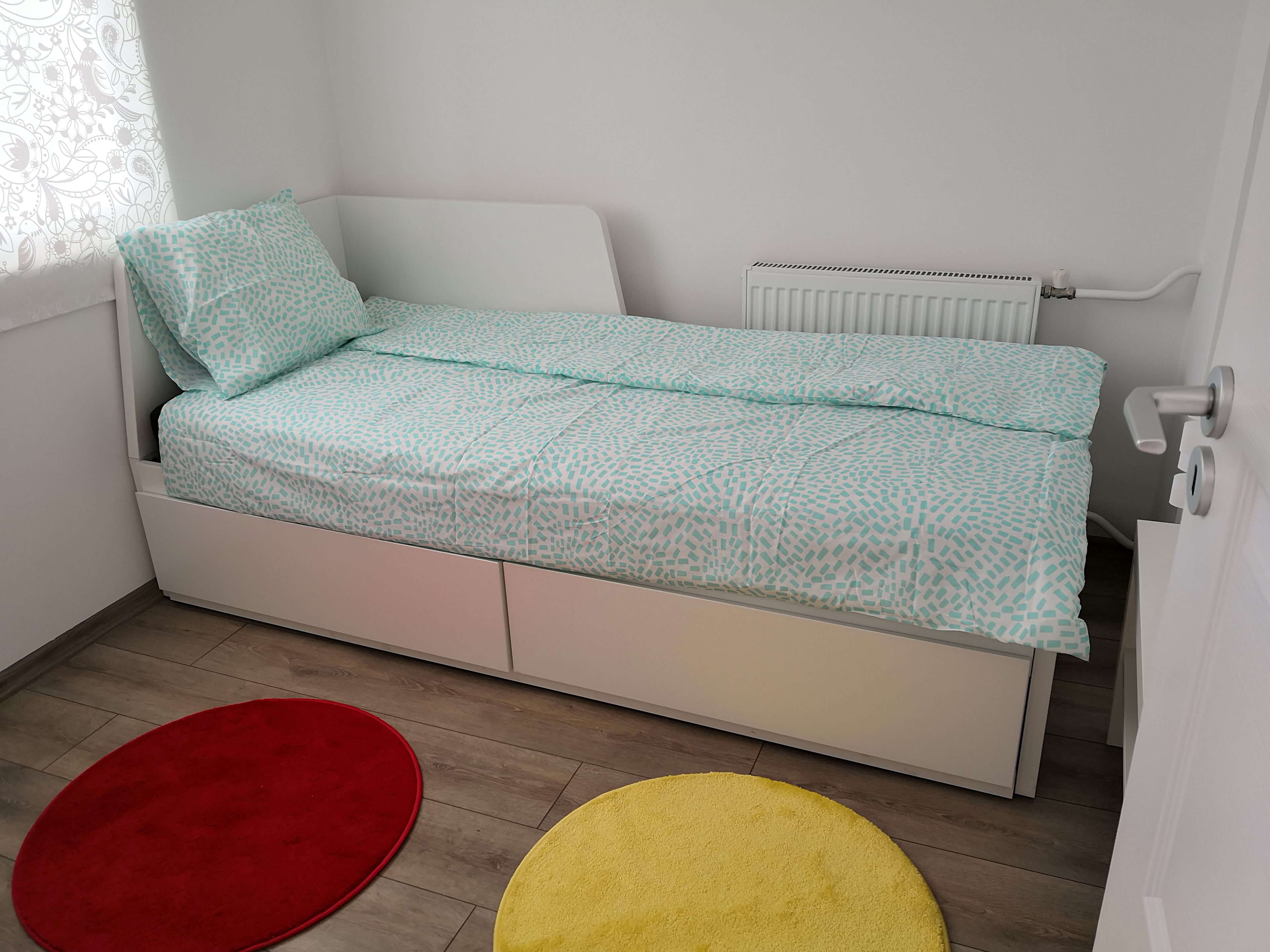 A small, white, single-bed room with a patterned bedspread, a red and a yellow rug, and a radiator on the wall.
