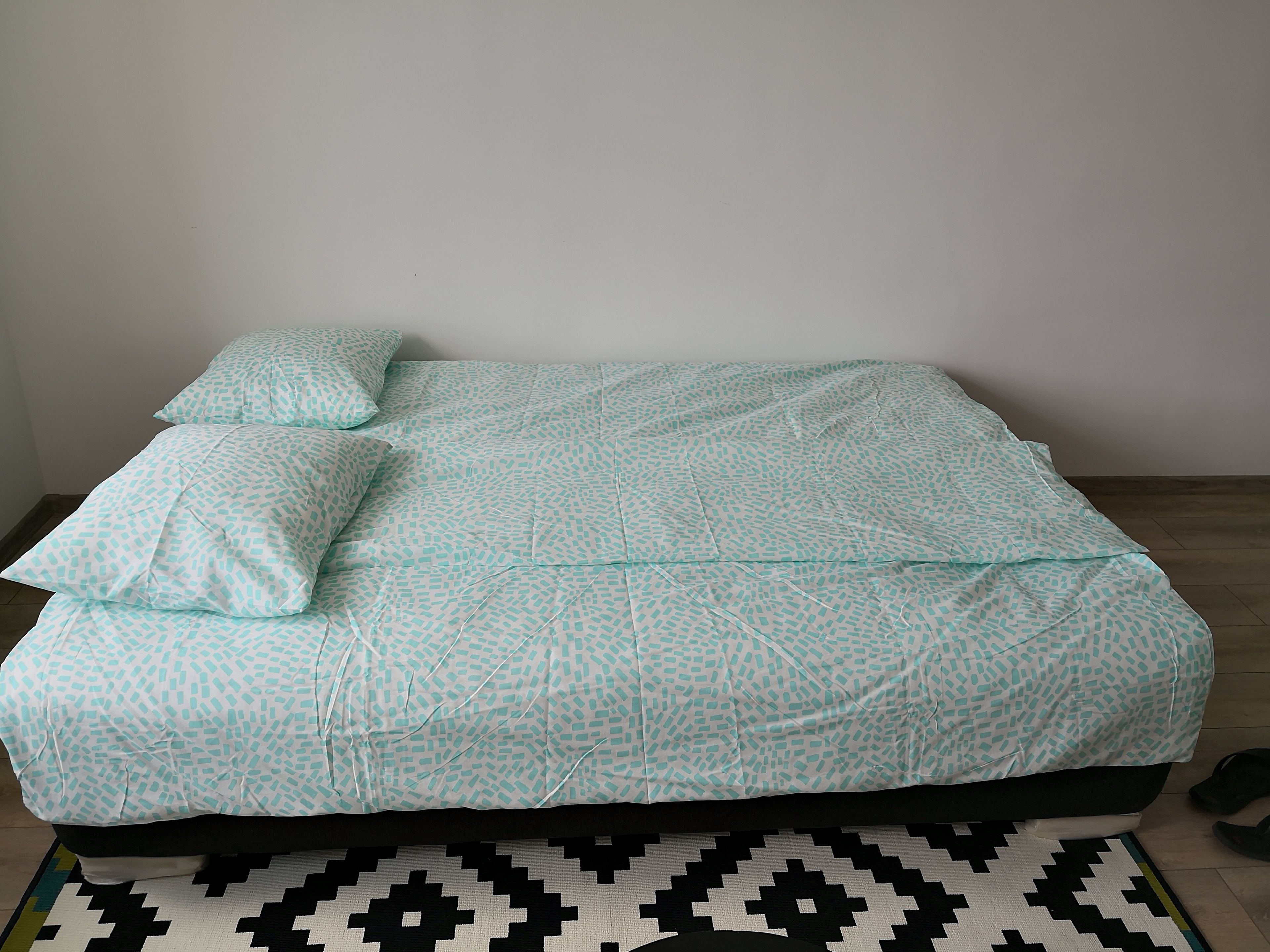 Ein Bett mit einem gemusterten Überwurf und Kissen, einem schwarz-weißen Teppich und einer schlichten Wand.