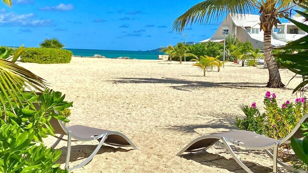 Ein Strand mit zwei Liegestühlen, einer Palme und einem Gebäude im Hintergrund.