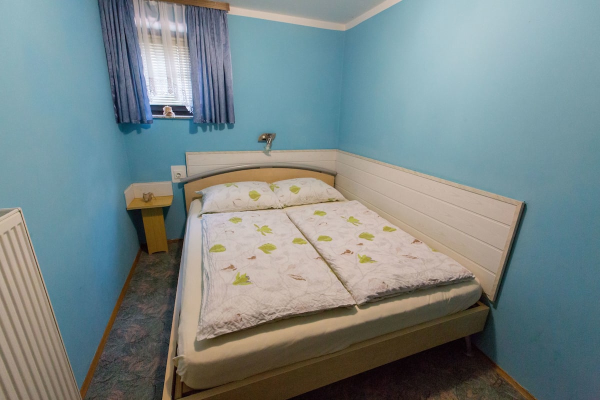 Una habitación pequeña con cama individual, cabecera de madera, mesita de noche y ventana con cortinas.