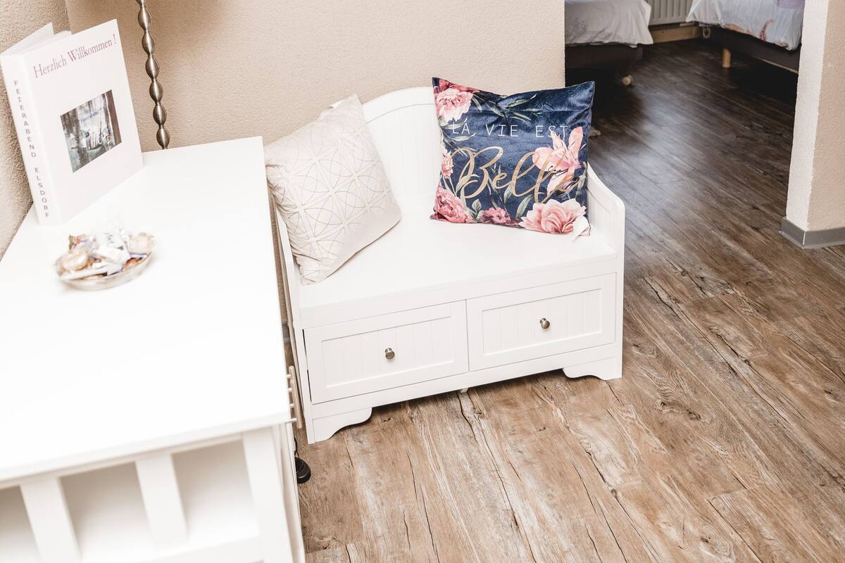 Een gezellig slaapkamer met een houten vloer, een witte nachtkastje met een lamp, een bloemenkussen en een ingelijst welkomstkaart.