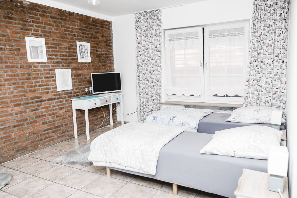 Een slaapkamer met een bakstenen muur, een bed met een grijze matras en witte beddengoed, een witte nachtkastje met een flatscreen televisie en een raam met gestreepte gordijnen.