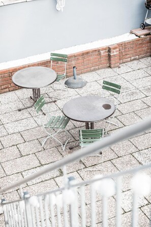 Un patio con dos mesas redondas y cuatro sillas verdes, un muro de ladrillo y una barandilla blanca.