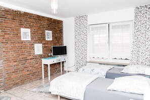 Un dormitorio con pared de ladrillo, un televisor sobre un escritorio blanco, una cama con colchón gris y cortinas estampadas.