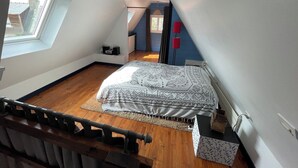 A bedroom with a sloped ceiling, a bed with patterned bedding, a radiator, a window, and a door leading to another room.
