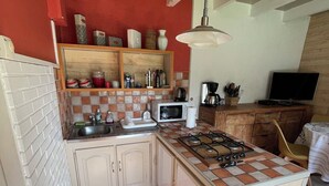 A compact kitchen with a microwave, gas stove, and a small sink. There is a wooden cabinet and a shelf with kitchen items.
