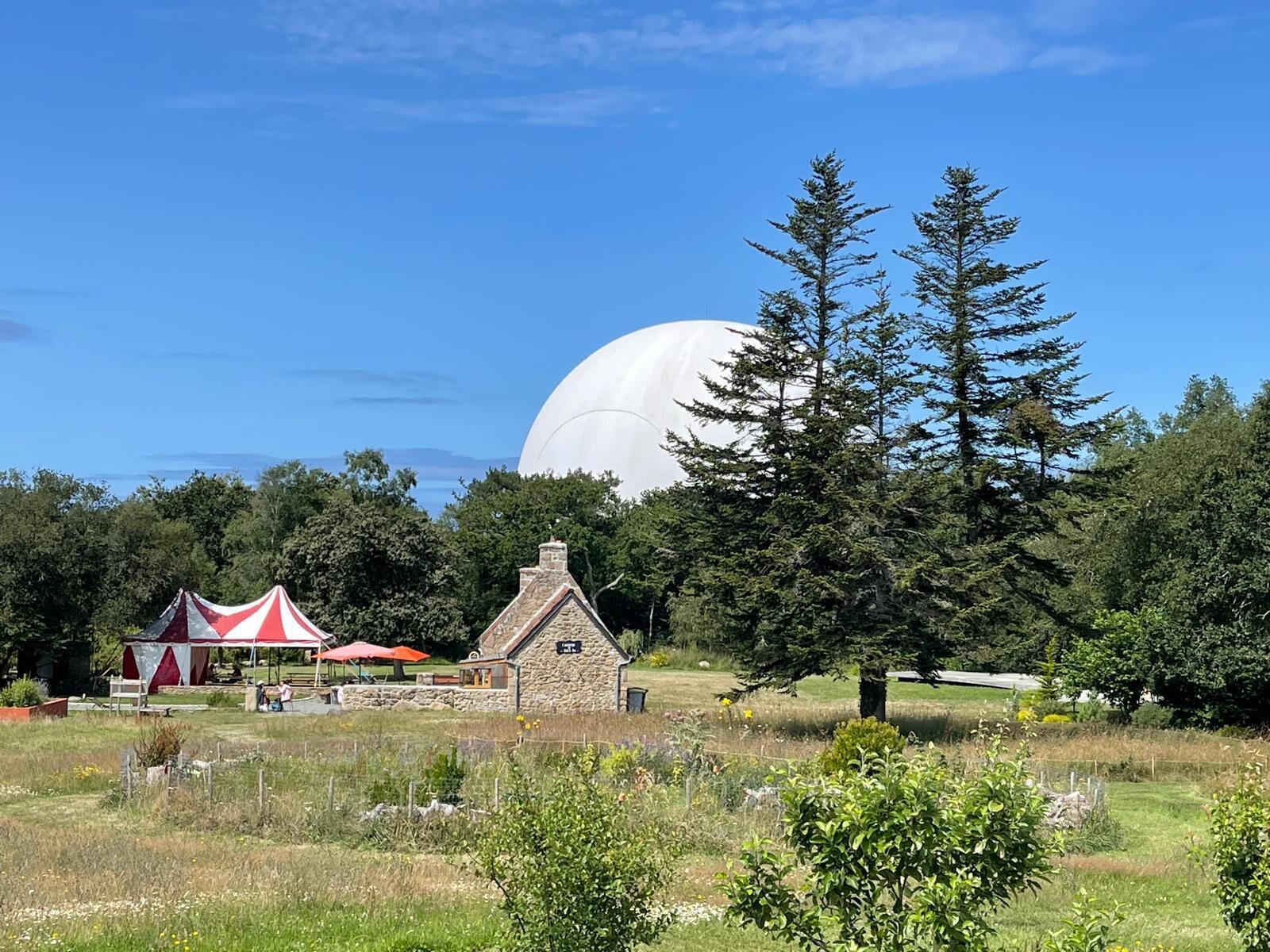 A rural landscape with a stone cottage, a red and white tent, and a large white dome structure.