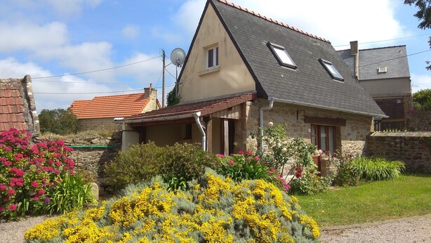 A stone house with a sloped roof, a small porch, and a garden with yellow flowers and pink shrubs.