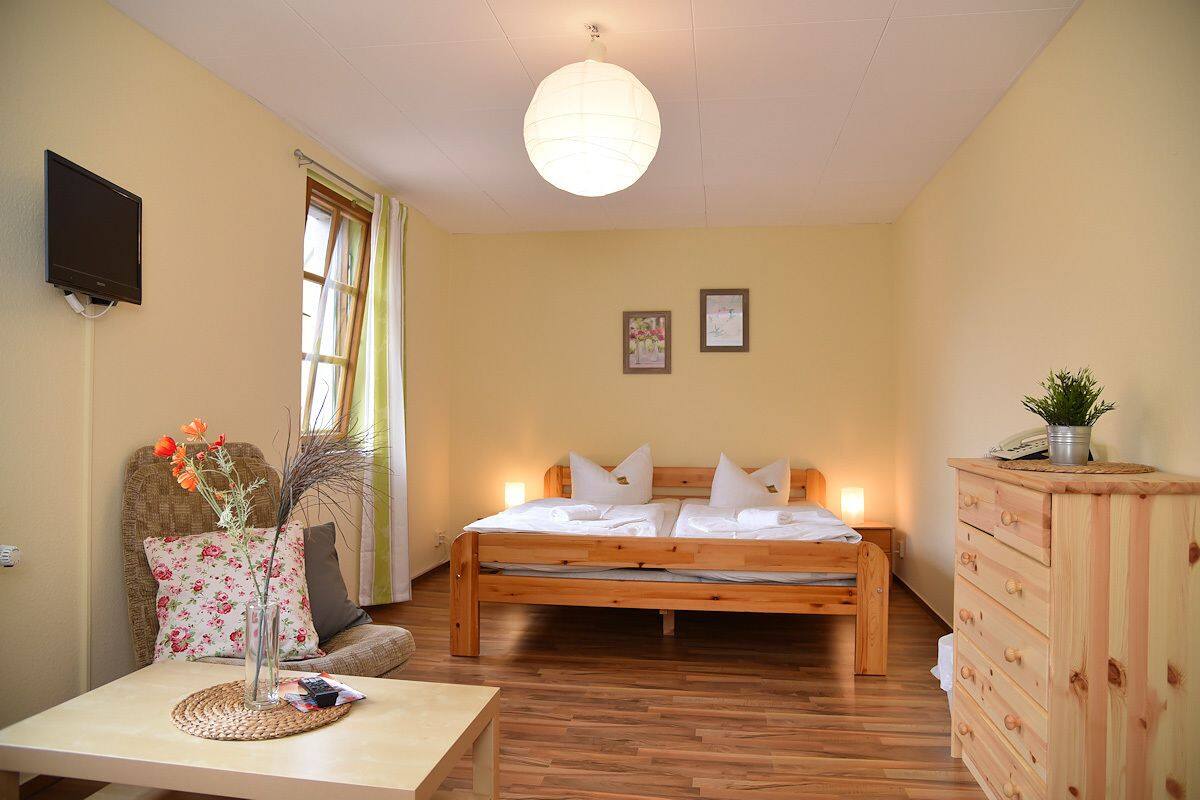 Un dormitorio con cama de madera, una mesita de noche con un florero, una silla, un aparador de madera y un televisor fijado a la pared.
