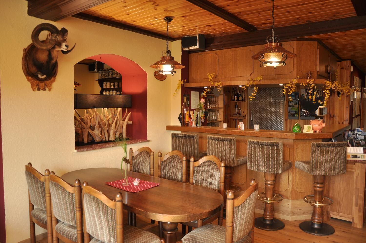 A dining area with a wooden table and chairs, a bar with stools, and a mounted ram head decoration.