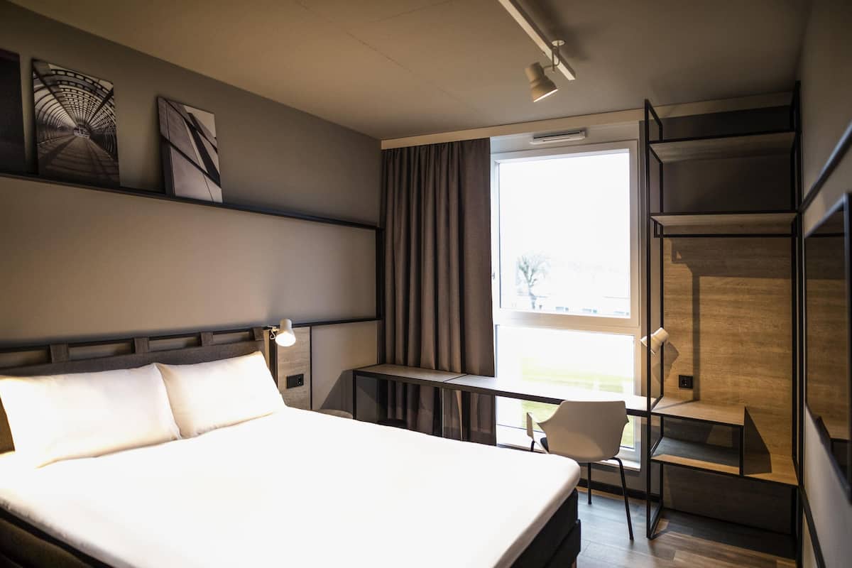 A modern hotel room with a large bed, a desk, a chair, and a view of the outdoors.