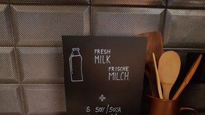 A kitchen counter with a chalkboard menu listing fresh milk options, a wooden spoon holder, and milk bottles. - Ibis Wels (Wels)