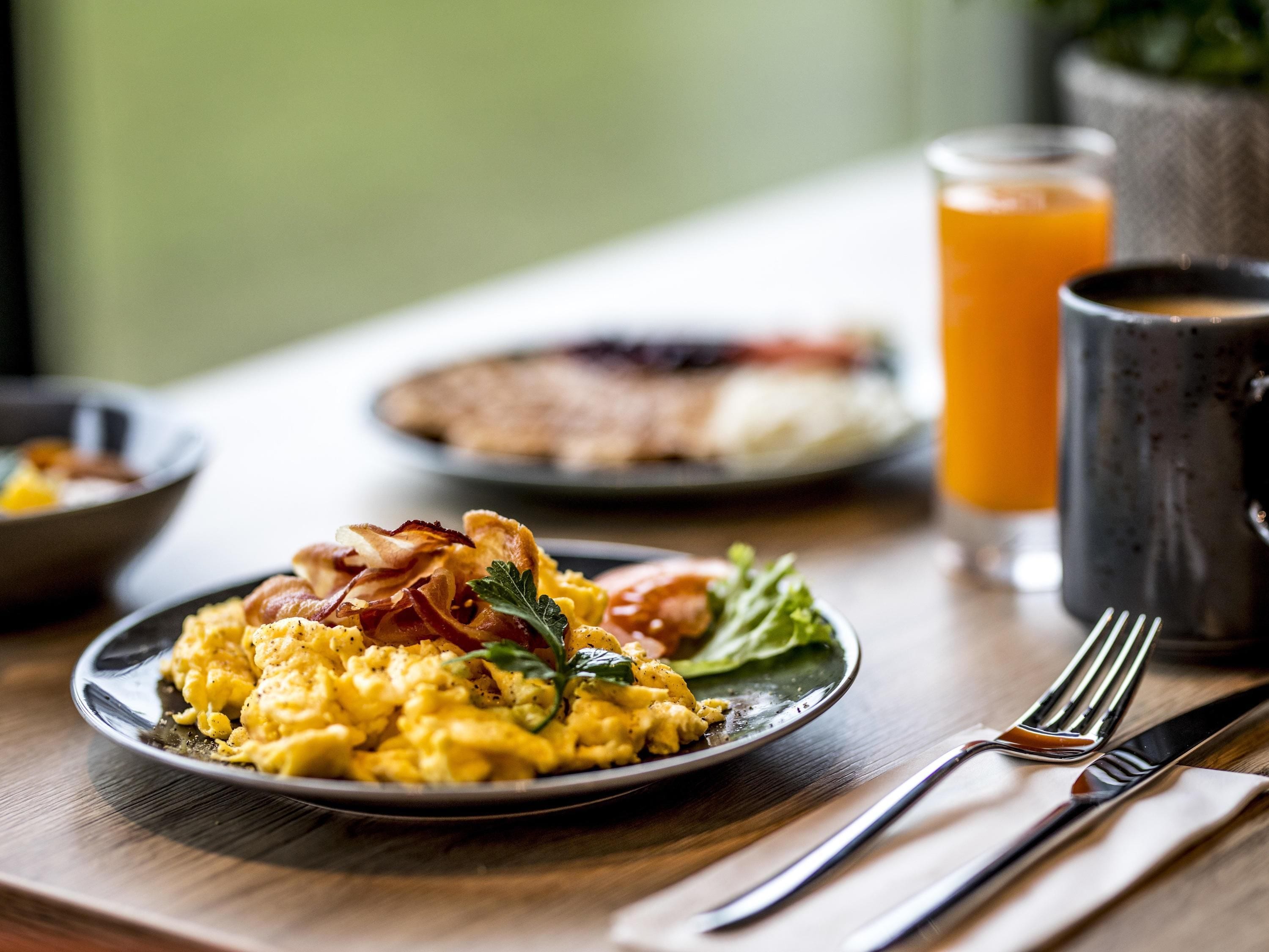Un plato de desayuno con huevos revueltos, tocino y decoración, un vaso de jugo de naranja y una taza sobre una mesa de madera.