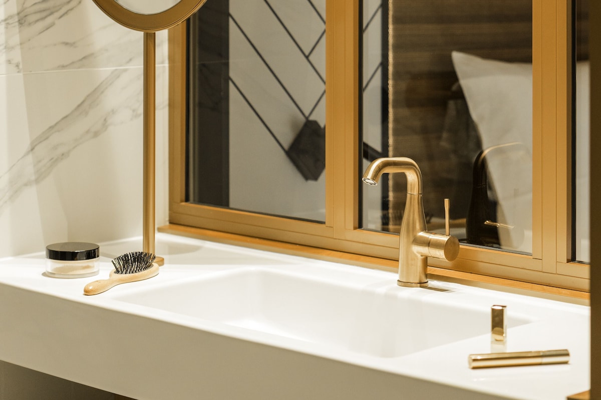 Een badkamer met een spiegel met gouden lijst, een witte wastafel en een raam met houten jaloezie.