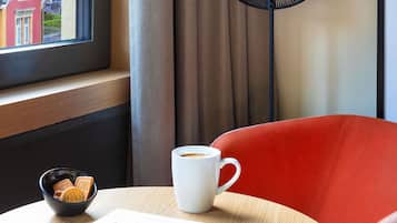 Hotelkamer met uitzicht uit het raam, een rode stoel, een witte mok, een schaal met koekjes en een boek op een houten tafel.