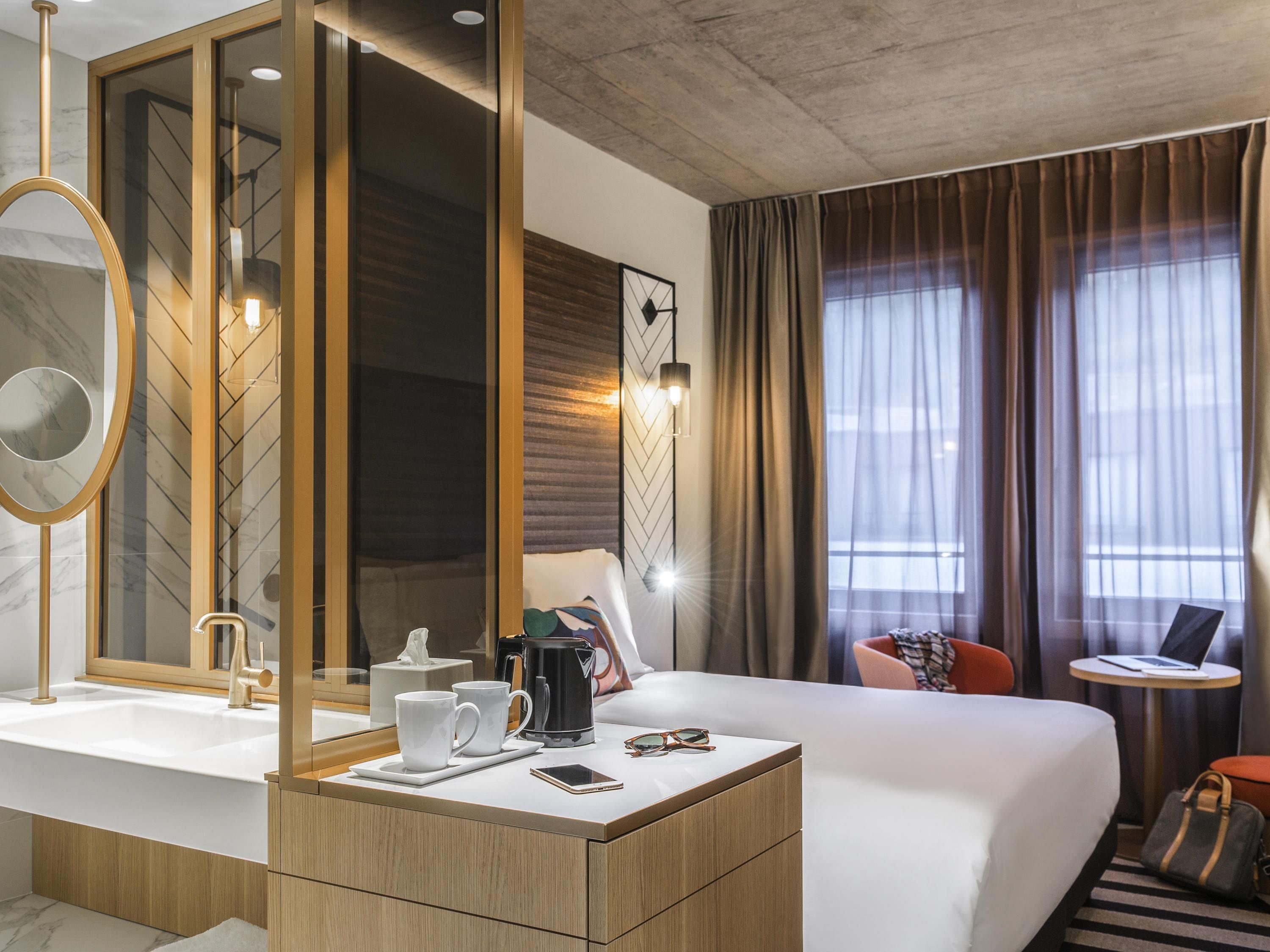 Habitación de hotel con una cama grande, una mesita de noche con teléfono y gafas, cafetera y vistas a la ciudad a través de amplias ventanas.