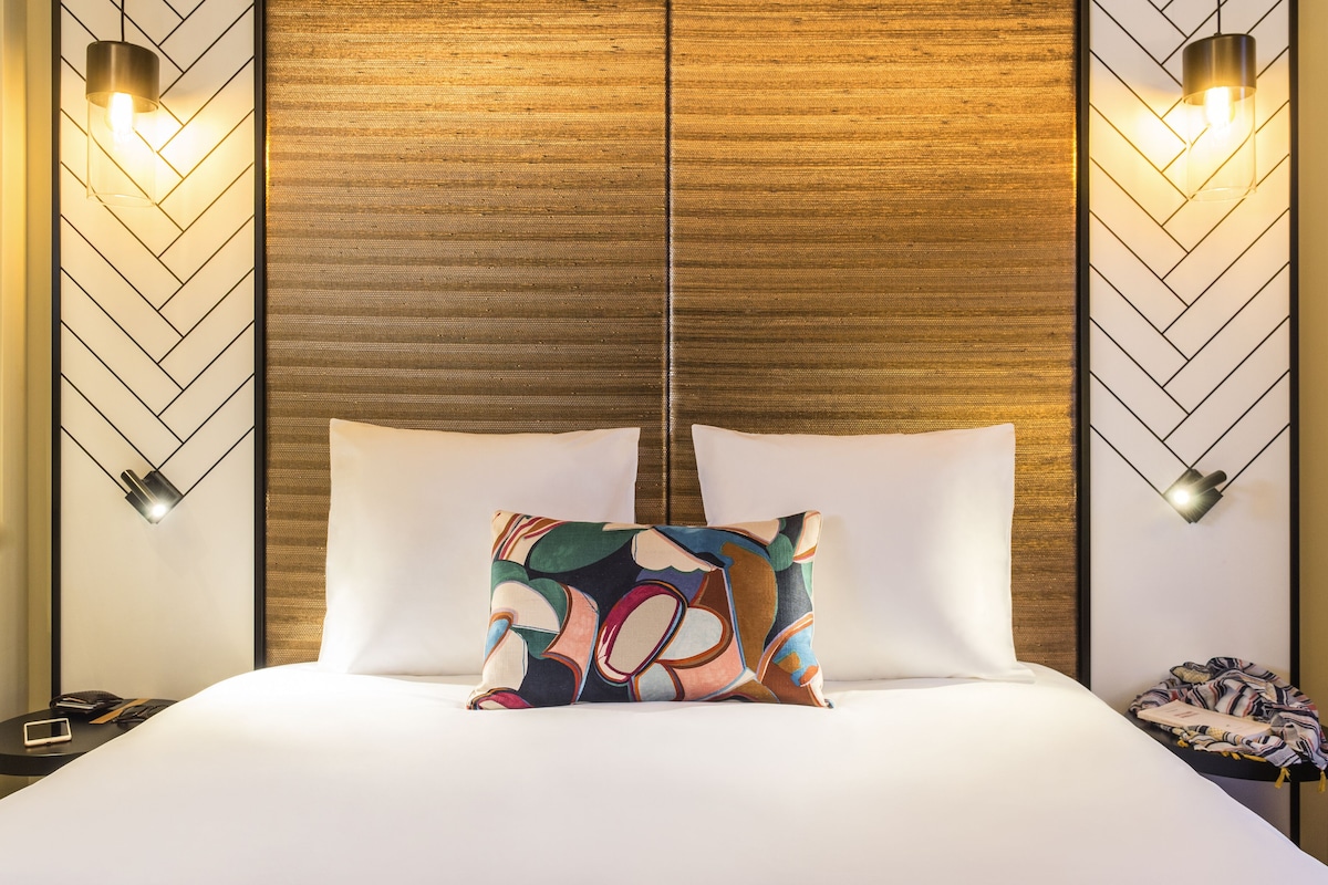 Een hotelkamer met een houten hoofdeinde, twee bedlampjes, een kleurrijk kussen en een net opgemaakt bed.