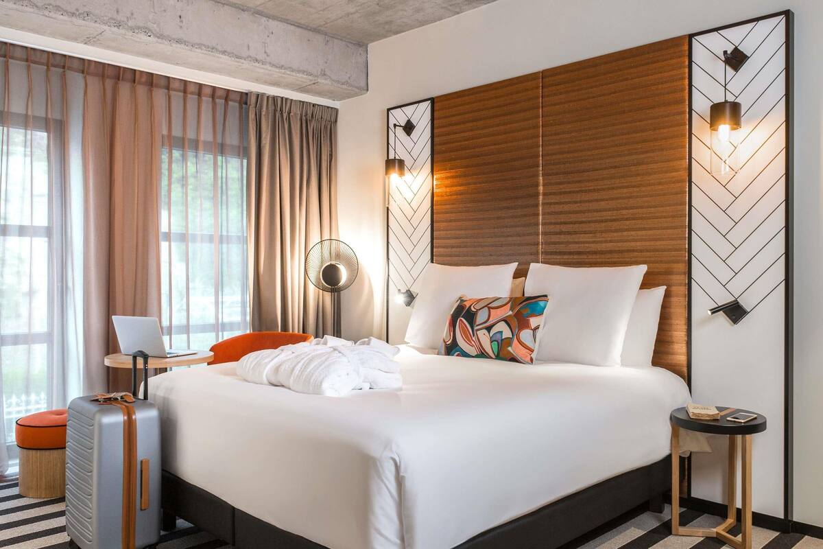 A hotel room with a bed, a suitcase, a lamp, and a view of the outdoors.