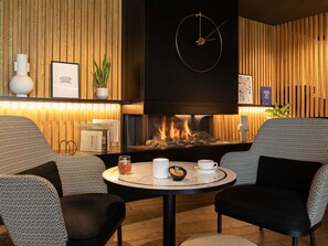 Un área de descanso acogedora con sillones estampados, una mesa redonda con bebidas, chimenea y un reloj de pared.