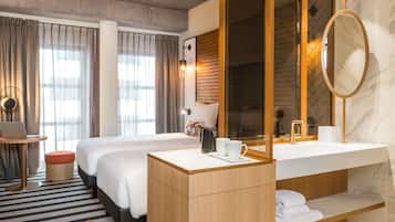 A hotel room with two beds, a desk with a laptop, a round mirror, a wooden vanity with a sink, and a large window with curtains.