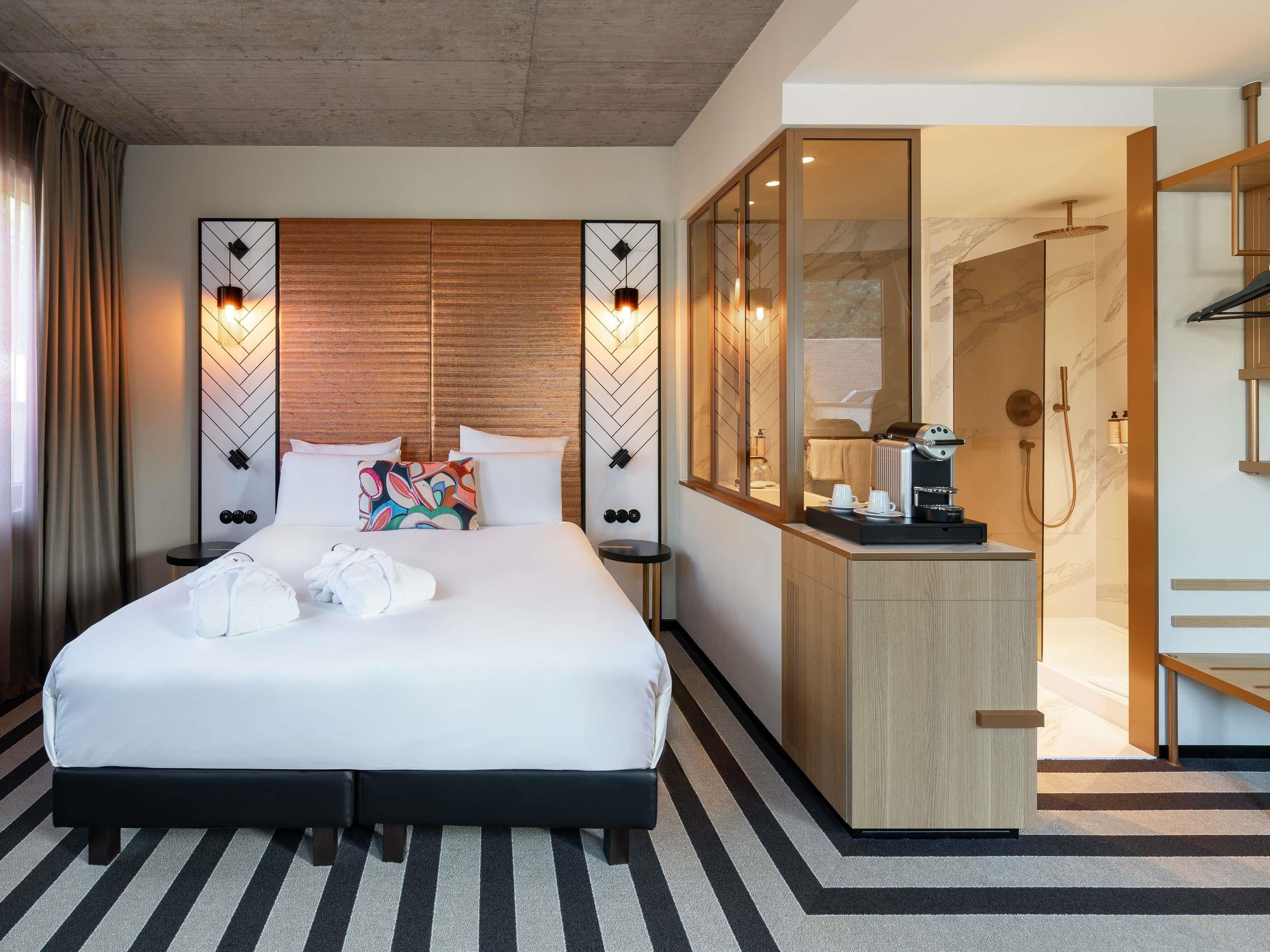 Habitación de hotel con una cama grande, una mesita de noche, un espejo, zona de ducha y una pequeña kitchenette.