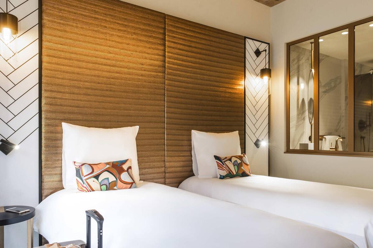A hotel room with two beds, a wooden headboard, a mirror, and a bedside table with a lamp.