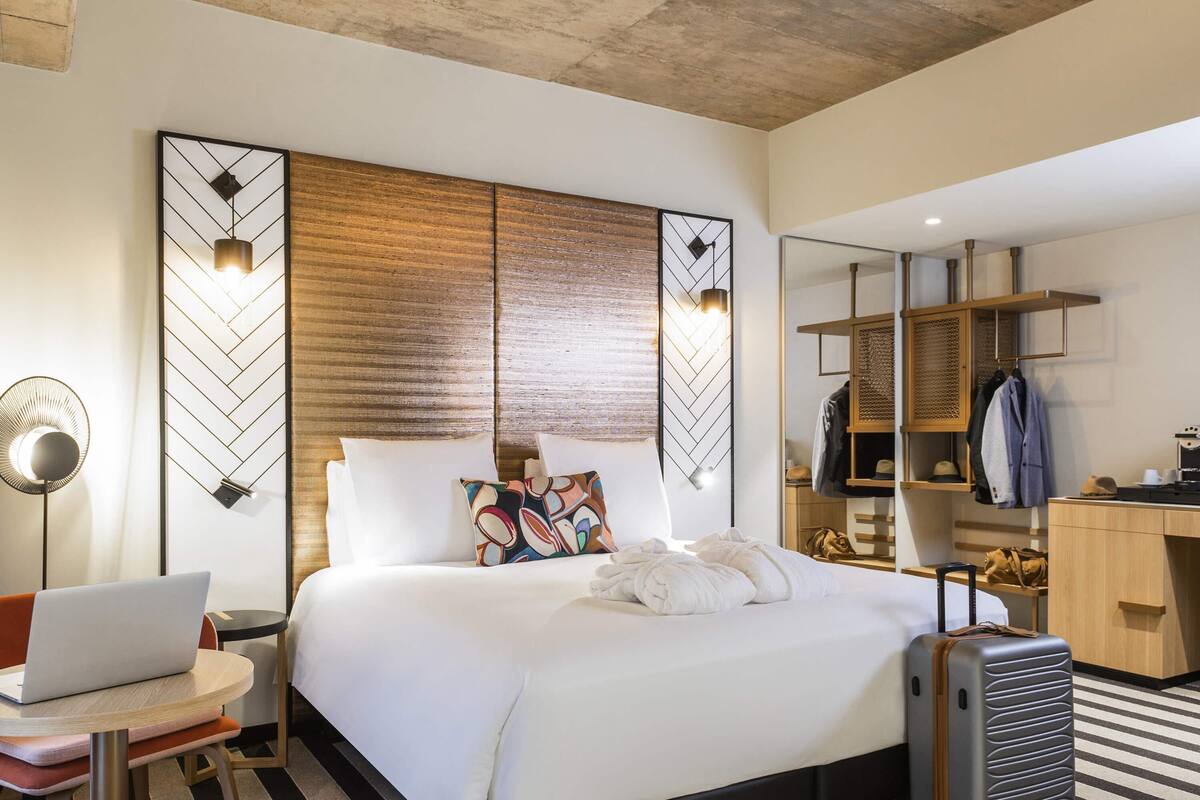 A hotel room with a bed, a desk with a laptop, a suitcase, and a wardrobe.