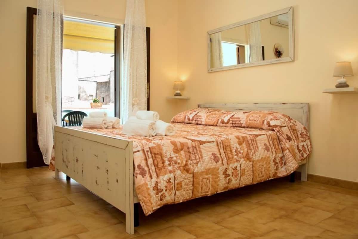 Ein Schlafzimmer mit Bett, Fenster mit Vorhängen, Spiegel und Nachttischlampe.
