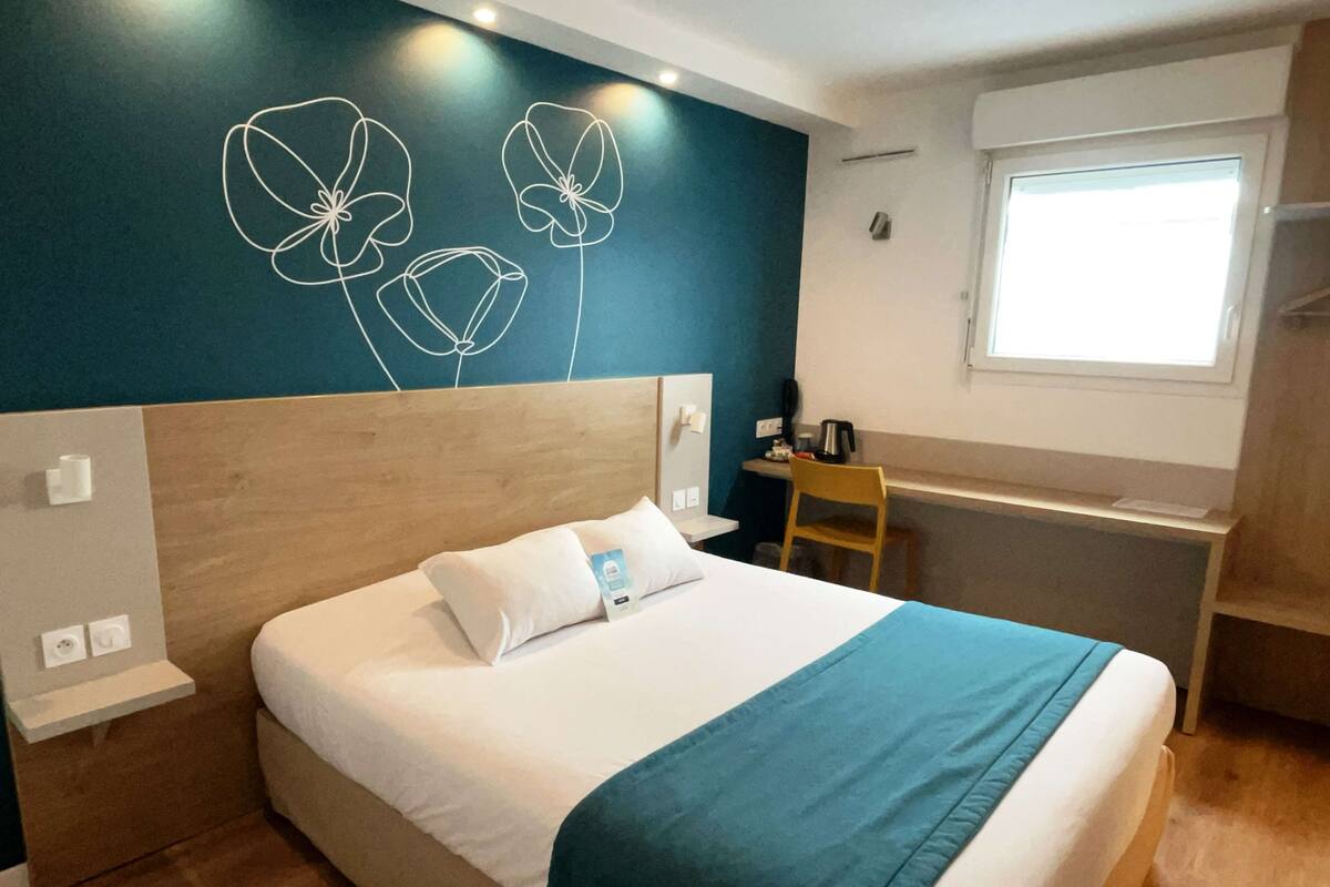 Une chambre d’hôtel comprenant un lit, un bureau, une chaise, une fenêtre et un mur orné d’un motif floral.