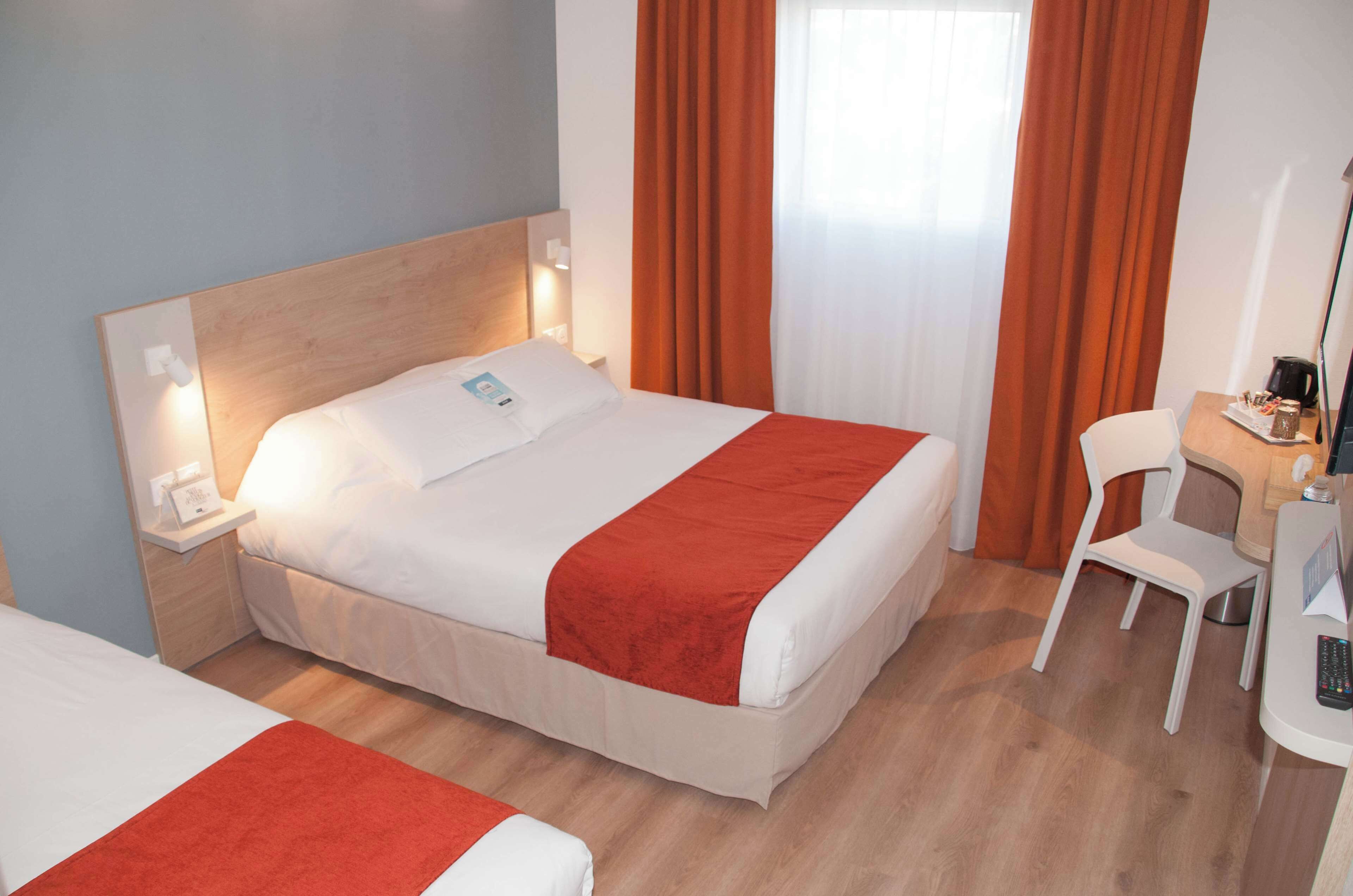 A hotel room with a bed, a desk, a chair, and a nightstand with a phone and a lamp.