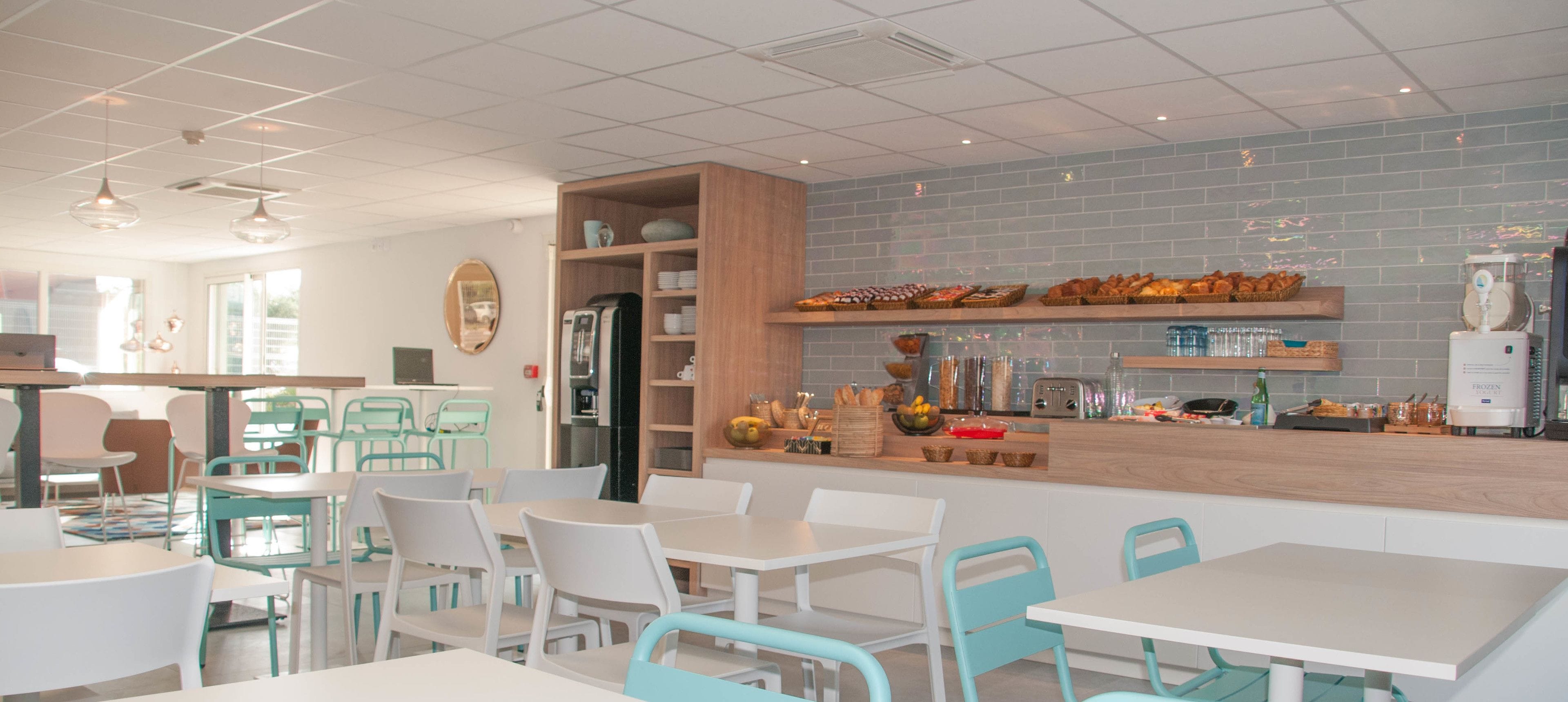 Un café avec des chaises blanches et turquoise, un comptoir en bois où sont exposés des plats, et un mur recouvert de carreaux.