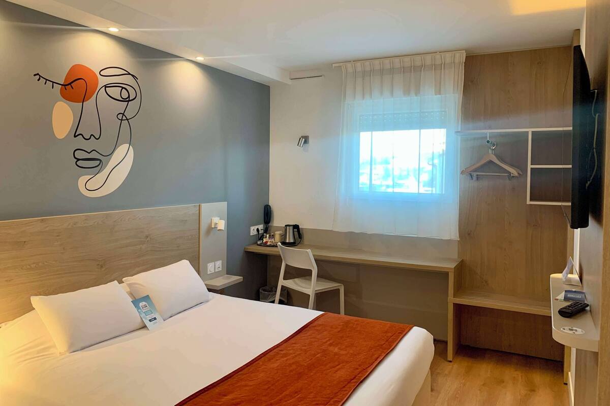 Une chambre d’hôtel avec un lit, un bureau, une chaise, une télévision et une fenêtre avec des rideaux.