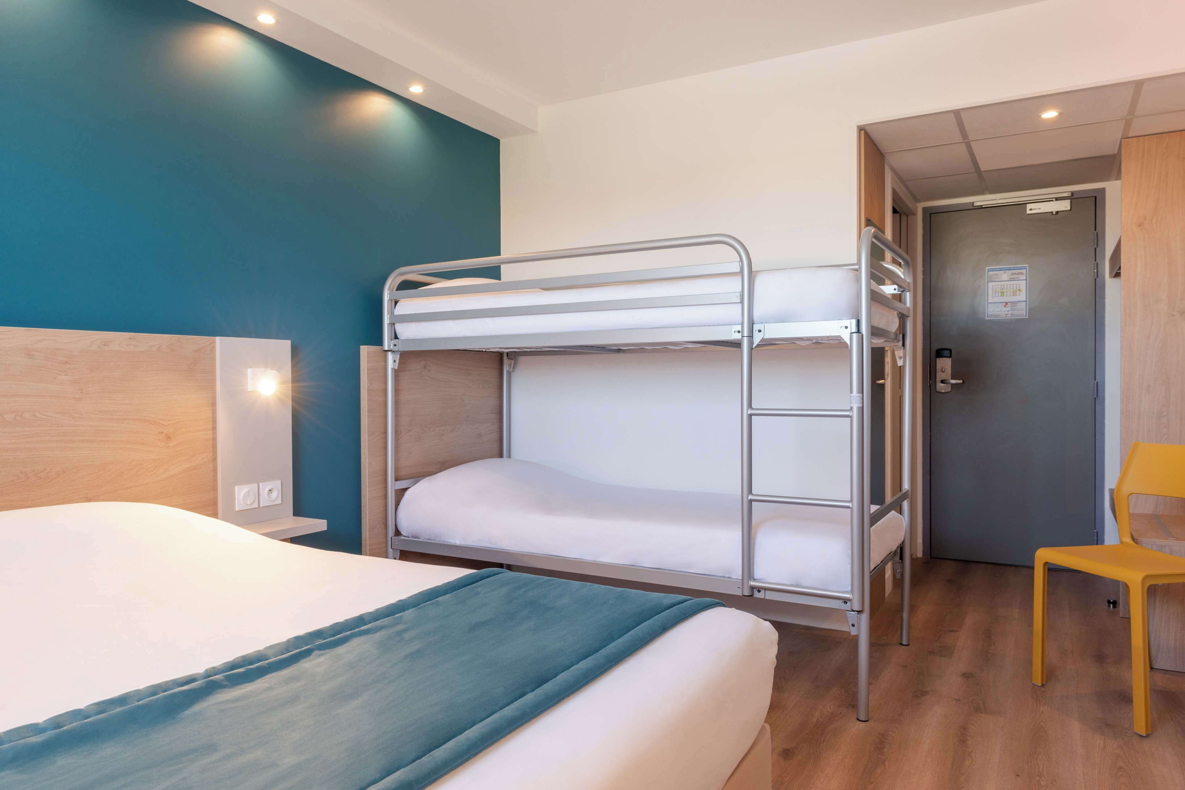 A hotel room with a bunk bed, a single bed, a wooden headboard, a yellow chair, and a door with a sign.